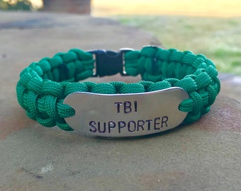 May include: Green paracord bracelet with a silver metal tag that reads "TBI Supporter".
