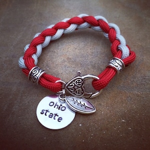 May include: Red and gray braided bracelet with a silver charm that says "Ohio State" and a football charm.