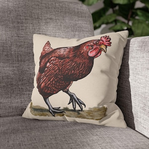 Puede incluir: Un cojín decorativo con una ilustración detallada de una gallina marrón. La gallina se representa caminando con la cabeza girada, mostrando su cresta y barbillas rojas. El cojín tiene un fondo neutro y está colocado sobre un sofá gris.