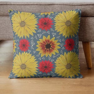 Puede incluir: Cojín decorativo cuadrado con un estampado floral. El cojín presenta grandes girasoles amarillos y flores rojas sobre un fondo azul. Pequeños corazones blancos están esparcidos alrededor de las flores. El cojín está sobre un suelo de madera.