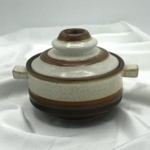 Puede incluir: Un cuenco de cerámica con tapa, cuerpo beige moteado y bandas marrones. La tapa tiene un diseño escalonado con la parte superior marrón. El cuenco tiene dos asas rectangulares pequeñas. El diseño es de estilo retro.