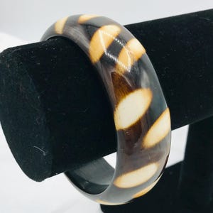 Puede incluir: Una pulsera de brazalete gruesa y redonda con un acabado brillante. La pulsera es de color marrón oscuro con acentos ovalados de color crema. La pulsera se exhibe sobre un soporte de terciopelo negro sobre un fondo blanco. Es un accesorio de moda.