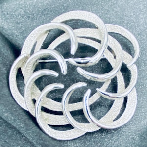 May include: A silver-toned brooch with a floral design. The brooch features interlocking, curved elements arranged in a circular pattern. The textured surface of the brooch reflects light, creating a subtle shimmer. A pin is attached to the back for secure fastening.