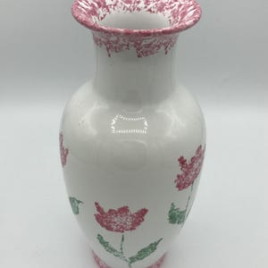 May include: White ceramic vase with a flared rim and a rounded body. The rim and base feature a speckled pink pattern. Pink floral designs with green leaves are painted on the vase, creating a charming, decorative piece.