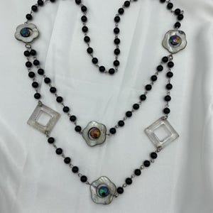 Può includere: Una collana a più strati con perline nere ed elementi decorativi. La collana include ciondoli a forma di fiore con centri iridescenti e ciondoli quadrati e trasparenti. Le perline sono infilate su una catena color argento.