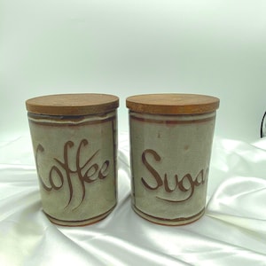May include: Two cylindrical ceramic canisters with light grey speckled glaze and brown wooden lids. The left canister features the word Coffee in brown script, while the right canister features the word Sugar in brown script, both with brown trim.