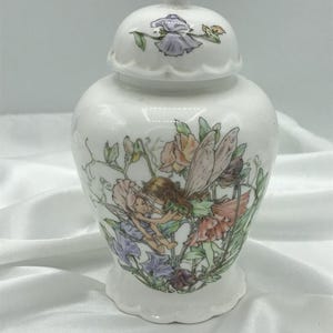 May include: A white porcelain jar with a lid, decorated with a floral and fairy design. The jar has a rounded body and a scalloped base. The lid is topped with a small finial. The artwork shows fairies amongst colourful flowers.