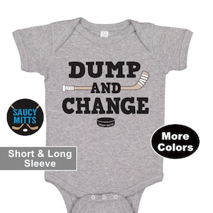 May include: A grey baby bodysuit with the text "DUMP AND CHANGE" in black. A hockey stick and puck graphic are included. The bodysuit has short sleeves and a three-snap closure. The brand logo "Saucy Mitts" is visible.