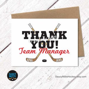 May include: A white card with the words "THANK YOU! Team Manager" in black and red, featuring crossed hockey sticks and a puck. The card is on a wooden surface with a brown envelope.