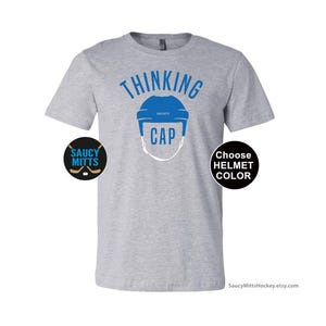 May include: Gray t-shirt with the text "Thinking Cap" and a blue hockey helmet graphic. The shirt also has two small logos, one that says "Saucy Mitts" and another that says "Choose Helmet Color".