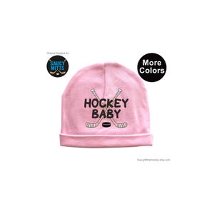 May include: Pink baby hat with the text "Hockey Baby" and a graphic of two crossed hockey sticks and a puck.