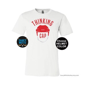 May include: White t-shirt with the text "Thinking Cap" in red. A red hockey helmet graphic is below the text. The text "Choose Helmet Color" is in a black circle on the right side of the shirt. The text "Saucy Mitts" is in a black circle on the left side of the shirt.