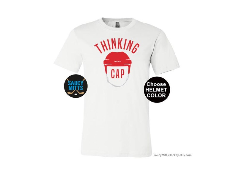 May include: White t-shirt with the text "Thinking Cap" in red. A red hockey helmet graphic is below the text. The text "Choose Helmet Color" is in a black circle on the right side of the shirt. The text "Saucy Mitts" is in a black circle on the left side of the shirt.