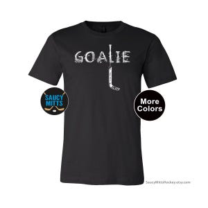Op de afbeelding: Zwart t-shirt met het woord "GOALIE" in het wit en een hockeystick-afbeelding. Het shirt heeft ook een "Saucy Mitts"-logo en een "Meer kleuren"-knop.