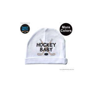 May include: White baby hat with the text "Hockey Baby" and a graphic of two crossed hockey sticks and a puck.