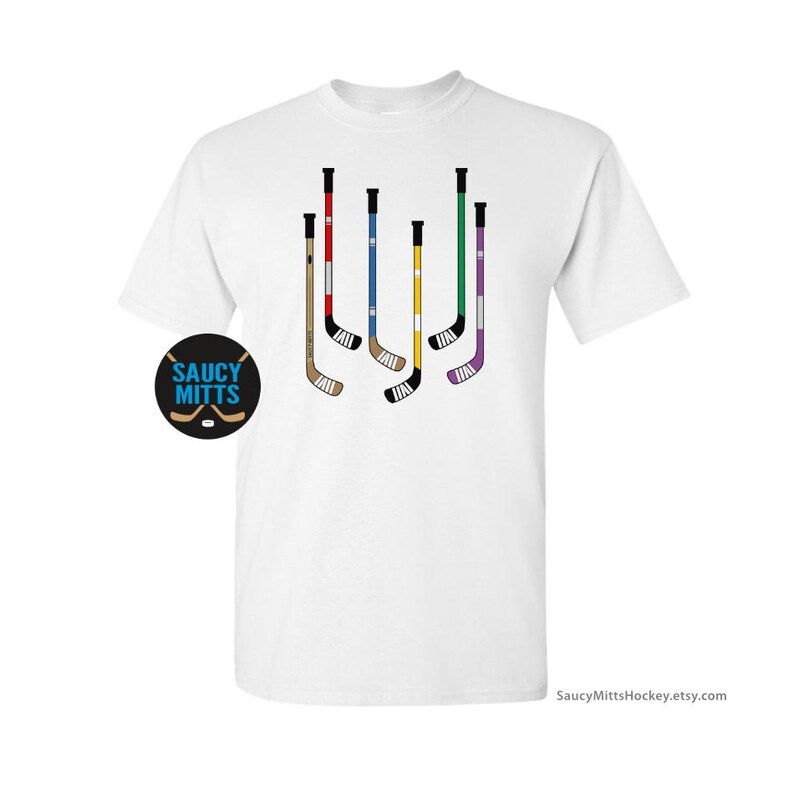 Op de afbeelding: Wit T-shirt met een afbeelding van zes hockeysticks in verschillende kleuren. De sticks zijn verticaal gerangschikt en hebben zwarte tape aan de onderkant. De tekst "Saucy Mitts" is in blauw op een witte achtergrond.