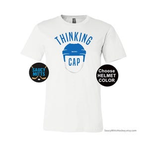 May include: White t-shirt with the text "Thinking Cap" in blue lettering. A blue hockey helmet graphic is above the text. The text "Choose Helmet Color" is in a black circle on the right side of the shirt. The text "Saucy Mitts" is in a black circle on the left side of the shirt.
