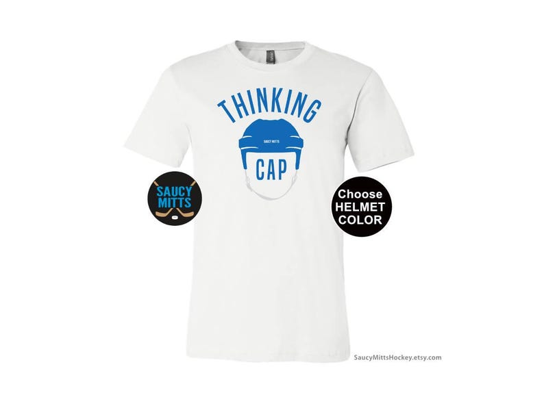 May include: White t-shirt with the text "Thinking Cap" in blue lettering. A blue hockey helmet graphic is above the text. The text "Choose Helmet Color" is in a black circle on the right side of the shirt. The text "Saucy Mitts" is in a black circle on the left side of the shirt.