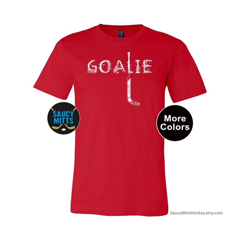 Op de afbeelding: Een rood t-shirt met het woord "GOALIE" in het wit, met een hockeystick-afbeelding. Het shirt heeft een "Saucy Mitts"-logo en een "More Colors"-knop.