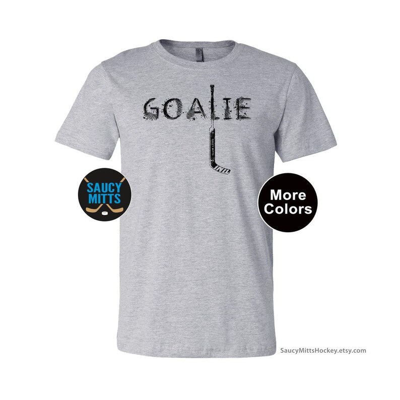 Op de afbeelding: Lichtgrijs t-shirt met het woord "GOALIE" in een distressed lettertype en een hockeystickafbeelding. Het shirt heeft ook een "Saucy Mitts" logo en een "Meer kleuren" knop.