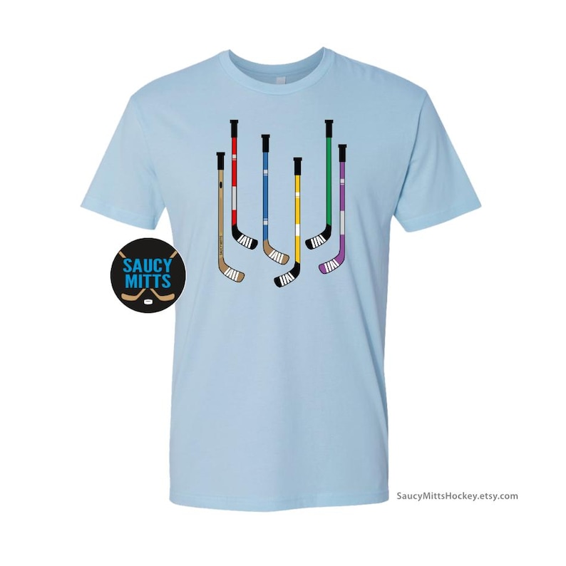 Op de afbeelding: Lichtblauw t-shirt met een afbeelding van zes hockeysticks in verschillende kleuren. De sticks zijn verticaal gerangschikt en hebben witte tape erop. De tekst "Saucy Mitts" staat in een cirkel linksonder op het shirt.