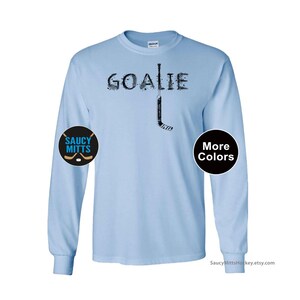 Op de afbeelding: Lichtblauw shirt met lange mouwen met het woord "GOALIE" in zwart, met een hockeystick-afbeelding. Het shirt heeft een rond "Saucy Mitts" logo op de linkermouw en een "More Colors" knop op de rechtermouw.