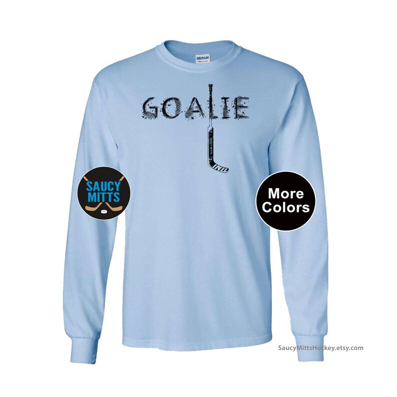 Op de afbeelding: Lichtblauw shirt met lange mouwen met het woord "GOALIE" in zwart, met een hockeystick-afbeelding. Het shirt heeft een rond "Saucy Mitts" logo op de linkermouw en een "More Colors" knop op de rechtermouw.