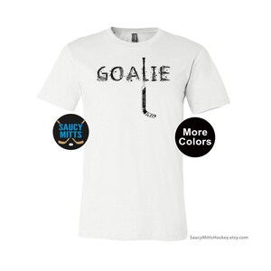 Op de afbeelding: Wit T-shirt met het woord "GOALIE" in een versleten lettertype en een hockeystickafbeelding. Een cirkelvormig logo met de tekst "SAUCY MITTS" en hockeysticks staat links. Een zwarte cirkel met "More Colors" staat rechts.