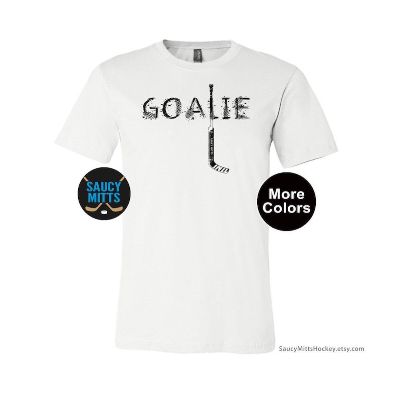 Op de afbeelding: Wit T-shirt met het woord "GOALIE" in een versleten lettertype en een hockeystickafbeelding. Een cirkelvormig logo met de tekst "SAUCY MITTS" en hockeysticks staat links. Een zwarte cirkel met "More Colors" staat rechts.