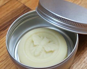May include: A round, off-white, solid bar of soap with a bee design embossed on the top. The soap is in a silver tin with a lid.