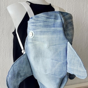 May include: A denim blue whale backpack with white straps. The whale has a white button on its side.