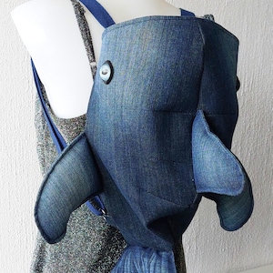 Puede incluir: Una mochila de mezclilla azul con forma de pez. La mochila tiene una cabeza grande, dos aletas y una cola. La mochila la lleva puesta una persona.