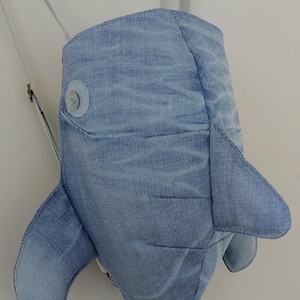 Puede incluir: Una mochila de ballena de mezclilla azul con un ojo de botón blanco. La mochila tiene una cola larga y dos aletas.