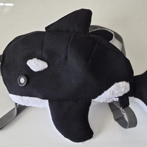 Puede incluir: Una mochila con forma de orca en blanco y negro con aleta dorsal y aleta caudal. La mochila tiene un ojo de botón y detalles en blanco. Las correas son grises.