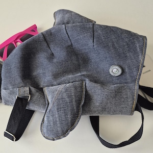 May include: A fish-shaped backpack made of dark gray denim fabric with a single silver button eye. The bag features black adjustable straps, a tail-shaped top section, and a side fin. Bright pink sunglasses are tucked near the top edge.
