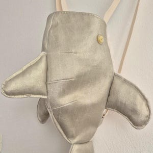 Può includere: Uno zaino a forma di squalo in argento metallizzato con due pinne laterali, una coda e un singolo bottone chiaro sul corpo superiore. La borsa include due sottili spallacci chiari fissati sulla parte superiore, creando un accessorio strutturato a forma di animale con una finitura scintillante.