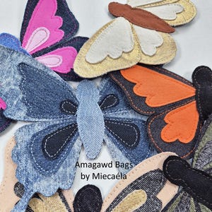 Op de afbeelding: Een verzameling stoffen vlinderapplicaties in diverse kleuren en texturen, waaronder denim, metallic goud, roze en oranje. De tekst Amagawd Bags by Miecaéla staat gedrukt in het midden van deze compositie van gelaagde textiele vlindervormen.