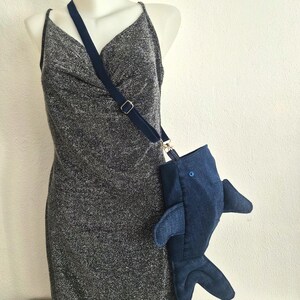 May include: A dark blue denim crossbody bag shaped like a shark, featuring side fins and a tail, attached to a dark blue adjustable fabric strap. The bag is displayed on a mannequin wearing a sleeveless, textured, dark gray metallic dress.
