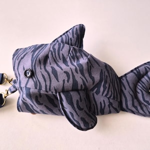 May include: A fish-shaped bag made of grey fabric with a dark grey tiger-stripe pattern. The bag features a dorsal fin, a tail fin, and a side fin. A small black button serves as an eye. Two dark blue fabric straps are attached with gold metal clips.