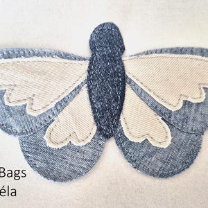 Op de afbeelding: Een vlindervormige stoffen applicatie met een lichaam van donkerblauwe denim en vleugels van lichtblauwe denim met crèmekleurige stoffen inzetstukken. De randen zijn afgewerkt met zichtbare stiksels. De tekst Amagawd Bags by Miecaéla is zwart gedrukt.