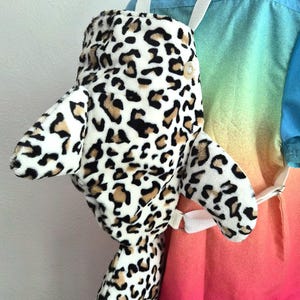 Può includere: Uno zaino in peluche a forma di delfino con un motivo leopardato bianco, nero e marrone chiaro. Lo zaino presenta spallacci in tessuto bianco, un bottone di colore chiaro sul lato e una consistenza morbida e soffice su tutto l'esterno.