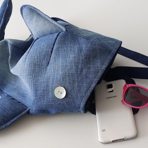 Puede incluir: Un bolso con forma de tiburón de mezclilla con cierre de botón, una correa azul marino, un teléfono Samsung blanco y gafas de sol rosas en el interior. El bolso está hecho de tela vaquera azul.