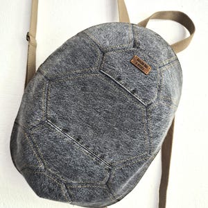 May include: A grey denim backpack with a geometric, football-inspired panel construction and tan webbing straps. A small rectangular brown leather label is stitched onto the upper right panel, featuring the text AMAGRID BAGS BY MITCHELLA.