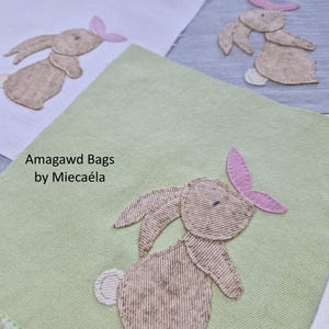 Puede incluir: Tres parches de tela cuadrados en blanco, gris y verde claro, cada uno con un conejo beige bordado con una oreja rosa y una cola blanca. El parche verde muestra el texto negro Amagawd Bags by Miecaéla. Cada conejo está cosido con un patrón de hilo texturizado.