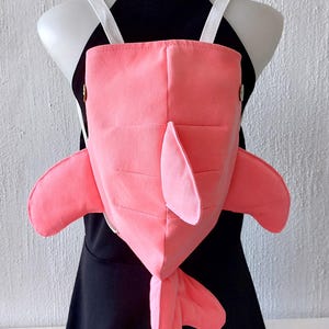 Puede incluir: Una mochila de color rosa en forma de ballena con correas blancas. La mochila tiene una cola y aletas.