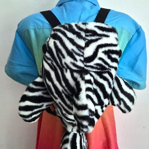 May include: A plush backpack featuring a black and white zebra stripe pattern. The bag has a soft, fuzzy texture, two black shoulder straps, and a distinct tail-like extension hanging from the bottom centre. The design includes two side fins.