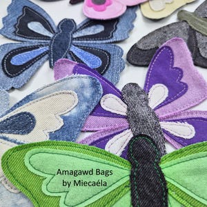 May include: A collection of fabric butterfly appliques in various colours including pink, blue, purple, green, and beige, featuring denim and cotton textures with stitched details. One green butterfly features the text Amagawd Bags by Miecaéla.