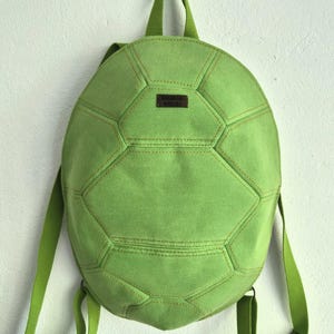 May include: A light green backpack shaped like a turtle shell with a textured fabric surface. The bag features stitched geometric panels resembling a shell pattern, adjustable shoulder straps, a top handle, and a small rectangular brown label.