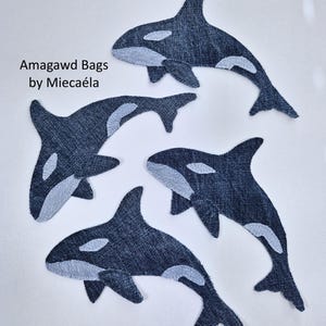 May include: Four orca whale patches crafted from dark blue denim fabric with light blue denim accents for the eye, dorsal, and side patches. The text Amagawd Bags by Miecaéla is printed in black on the left side of the image.
