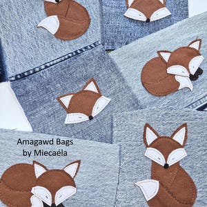 Può includere: Diversi pezzi di tessuto denim con applicazioni ricamate di volpi in marrone e bianco in varie pose. Alcune volpi sono sedute in posizione eretta mentre altre sono raggomitolate. Il testo Amagawd Bags by Miecaéla è stampato su una sezione.